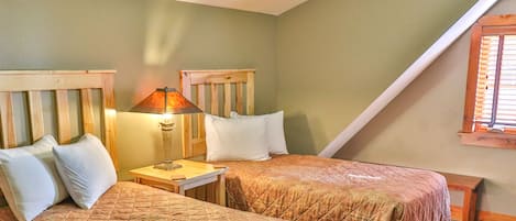 Una habitación con dos camas, cada una con cabeceros de madera y almohadas blancas. Un mesita de noche con lámpara se encuentra entre las camas.