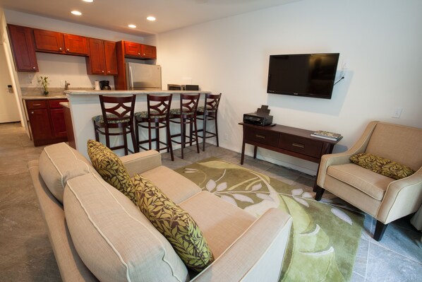A modern kitchen with wooden cabinets, a dining area with a sofa and chairs, and a flat-screen TV mounted on the wall. - 2 Bedroom Kauai Vacation Rental (Kapaa)