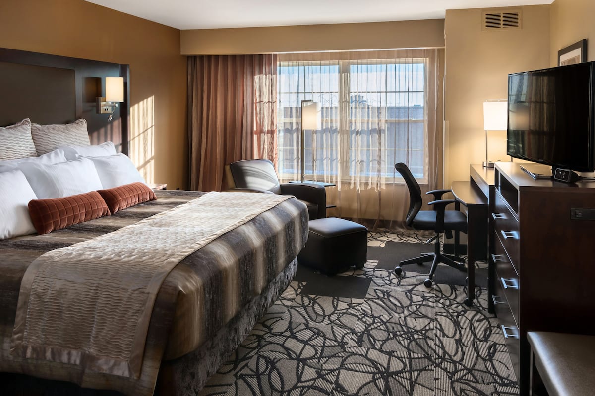 A hotel room with a large bed, a desk with a television, a chair, and a view of the city through large windows.