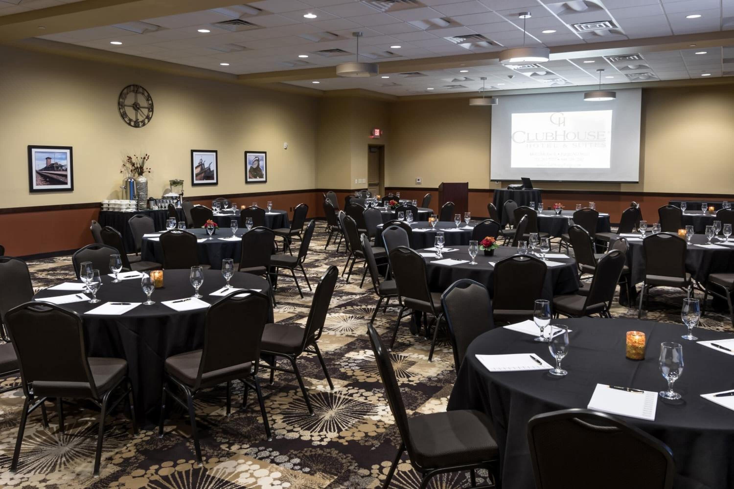 A conference room with multiple tables and chairs, a clock on the wall, and a projector screen displaying "Clubhouse".