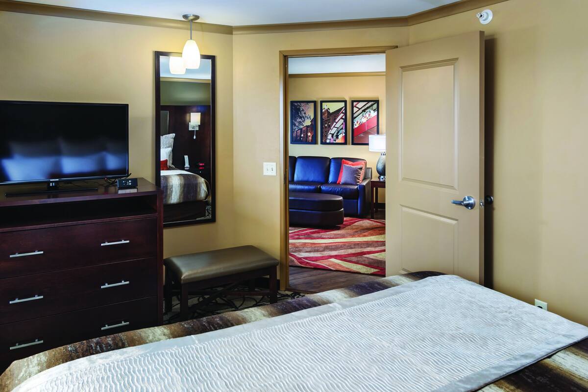 A hotel room with a bed, a TV, a dresser, a bench, and a door leading to another room.