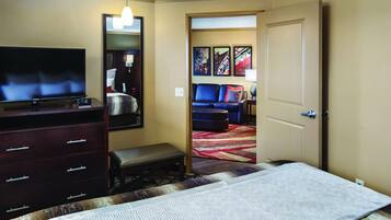 Quarto de hotel com cama, televisão, penteadeira, um banco e porta que dá para outro cômodo.