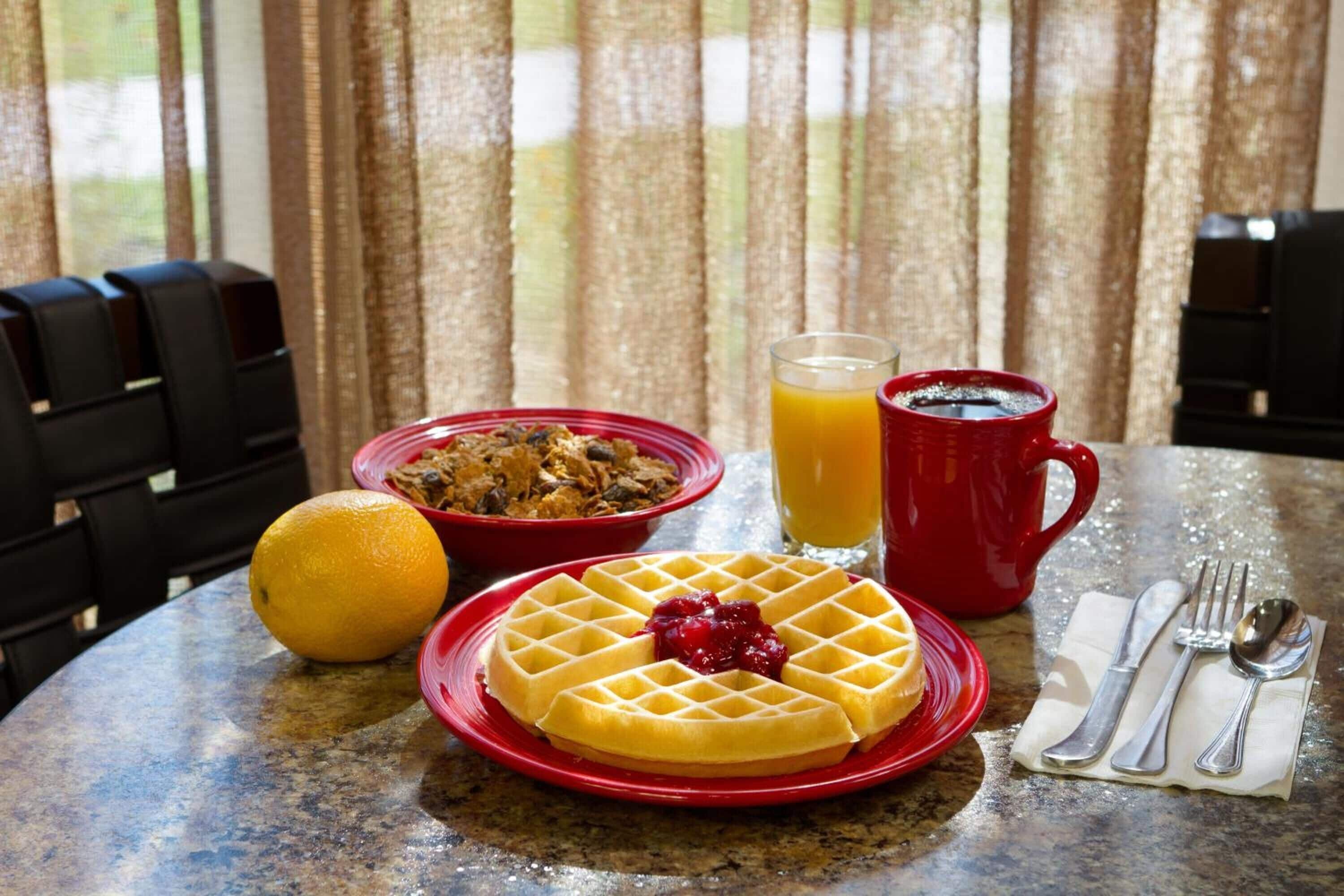 Una mesa de desayuno con waffles, zumo de naranja, café y un tazón de cereal.