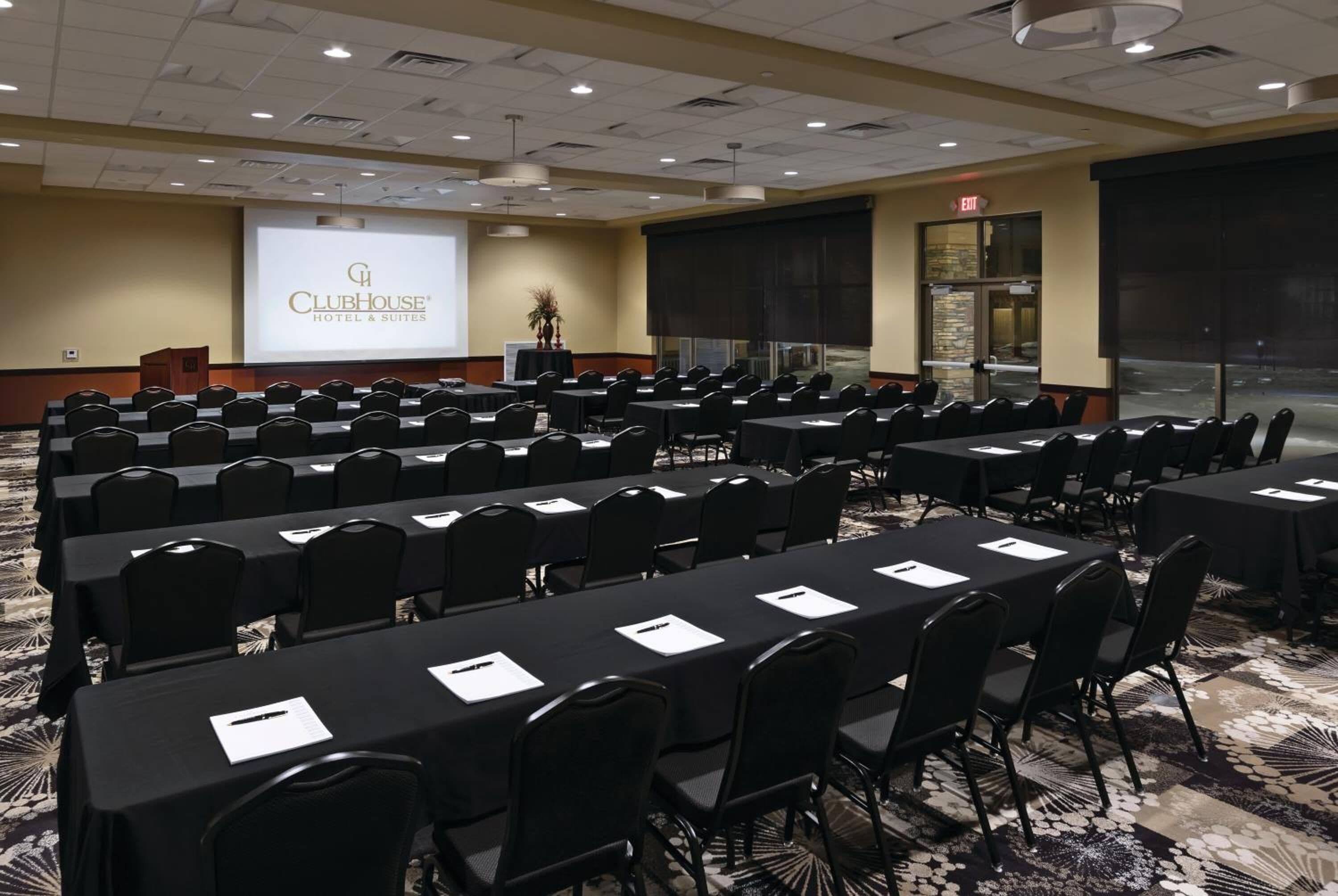 Une salle de conférence équipée de plusieurs tables et chaises, d’un pupitre et d’un écran de présentation affichant le nom « ClubHouse Hotel & Suites ».