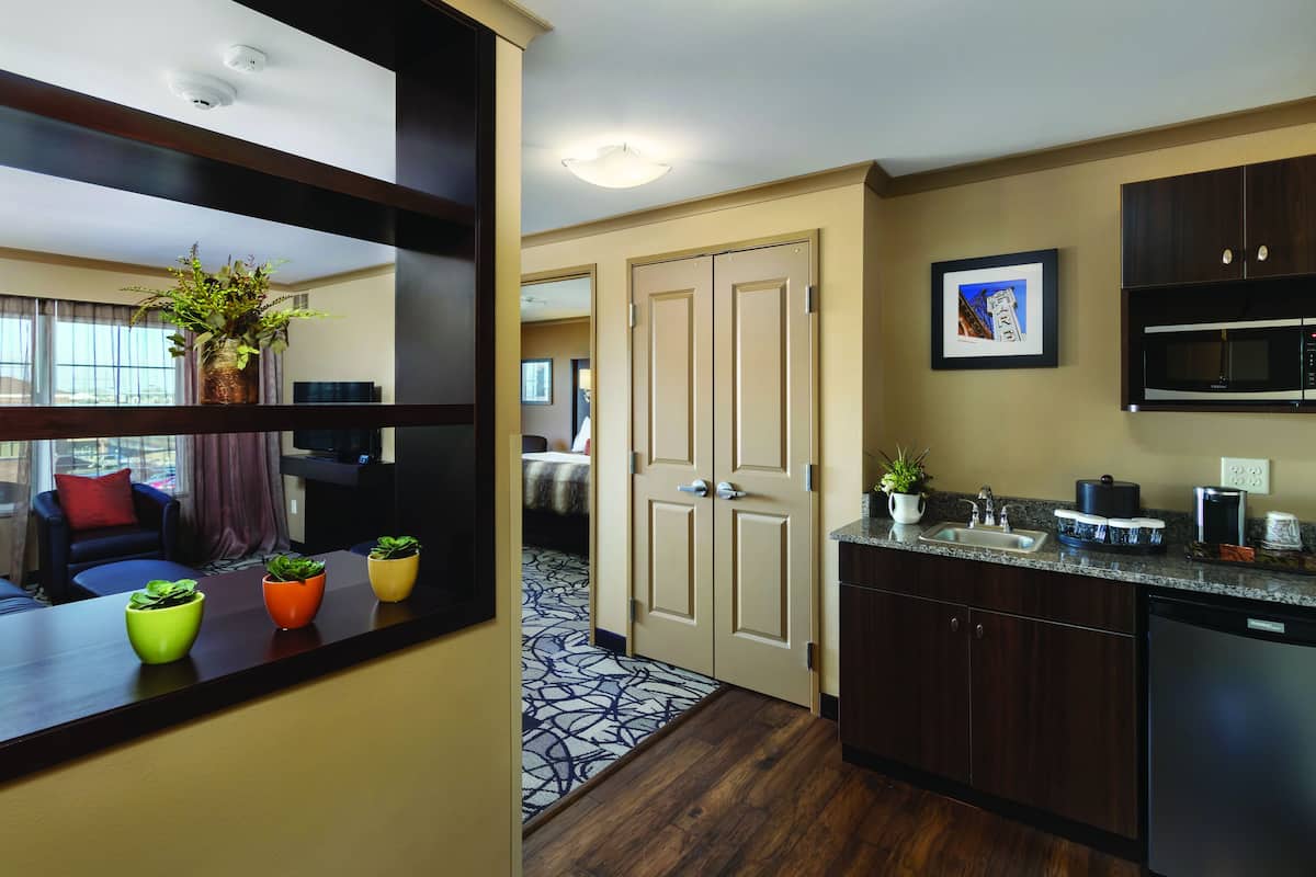 A modern hotel room with a kitchen area, a dining area with a table and chairs, and a hallway leading to a bedroom.