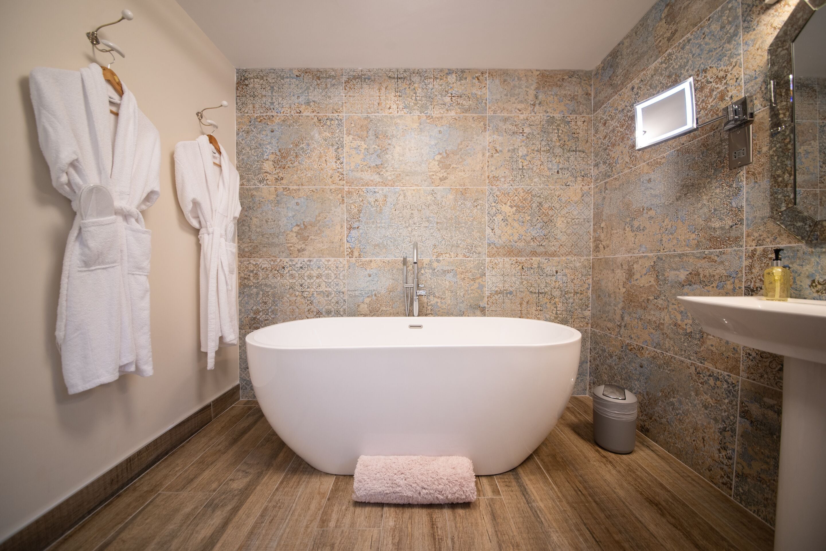 A modern bathroom with a freestanding bathtub, a wall-mounted towel rack with robes, a sink, and a trash bin.