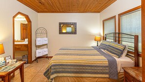 A hotel room with a wooden ceiling, a bed with striped bedding, a bedside table, a lamp, a mirror, and a window with blinds. - River Rock Inn (Mariposa)