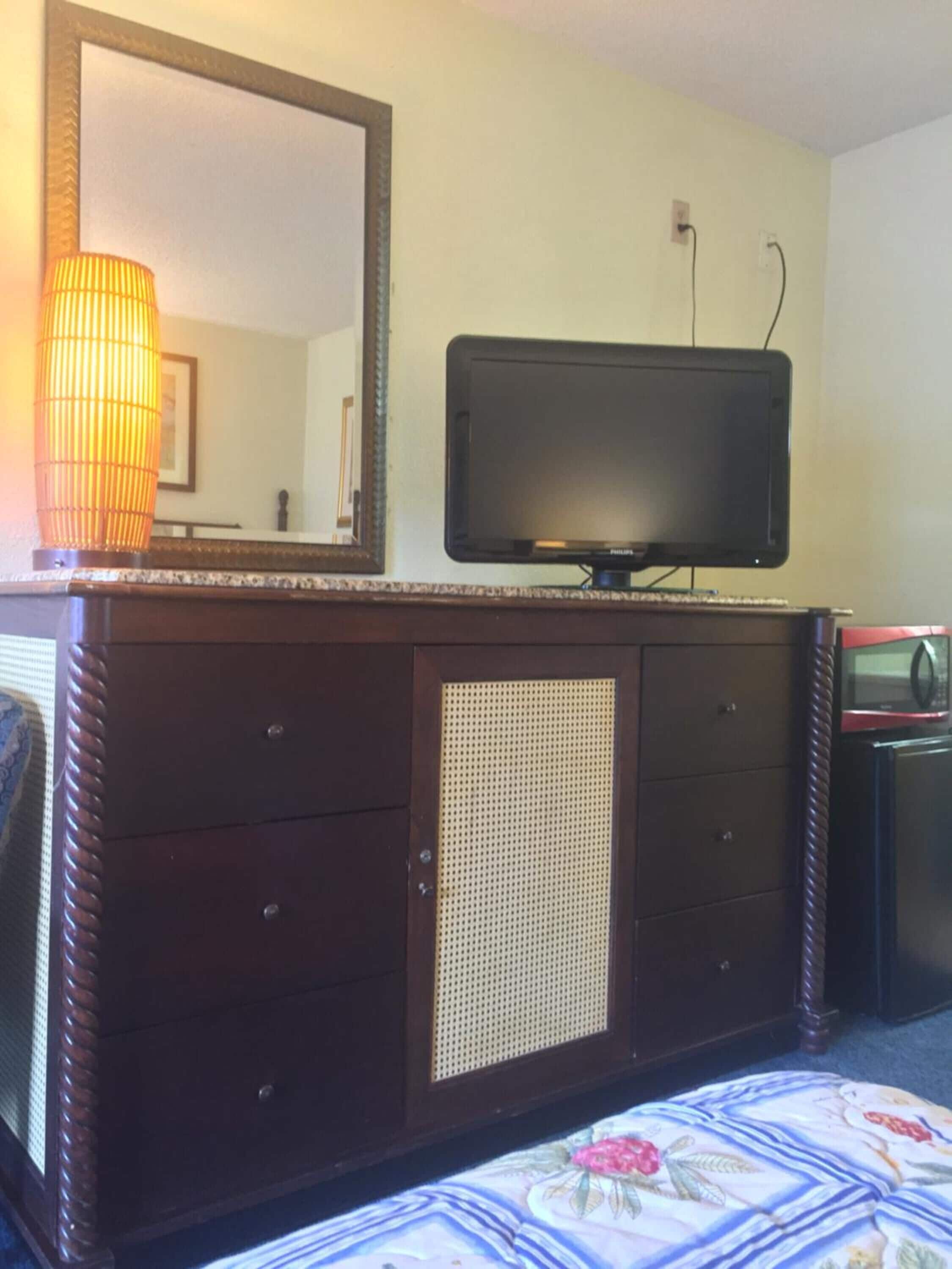 A hotel room with a large mirror, a flat-screen TV, a wooden dresser with drawers, a microwave, and a bed with a floral bedspread.