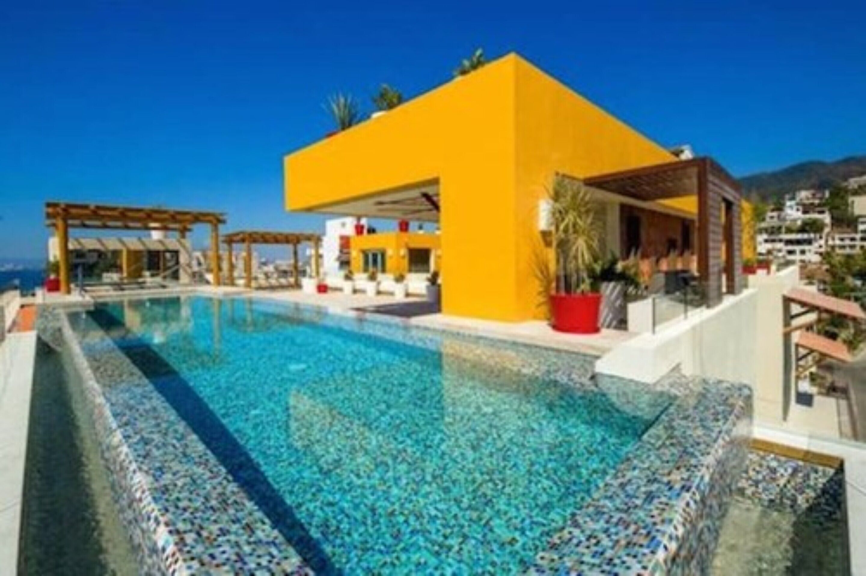 Una piscina con borde de azulejos, un edificio amarillo y un cielo azul despejado.