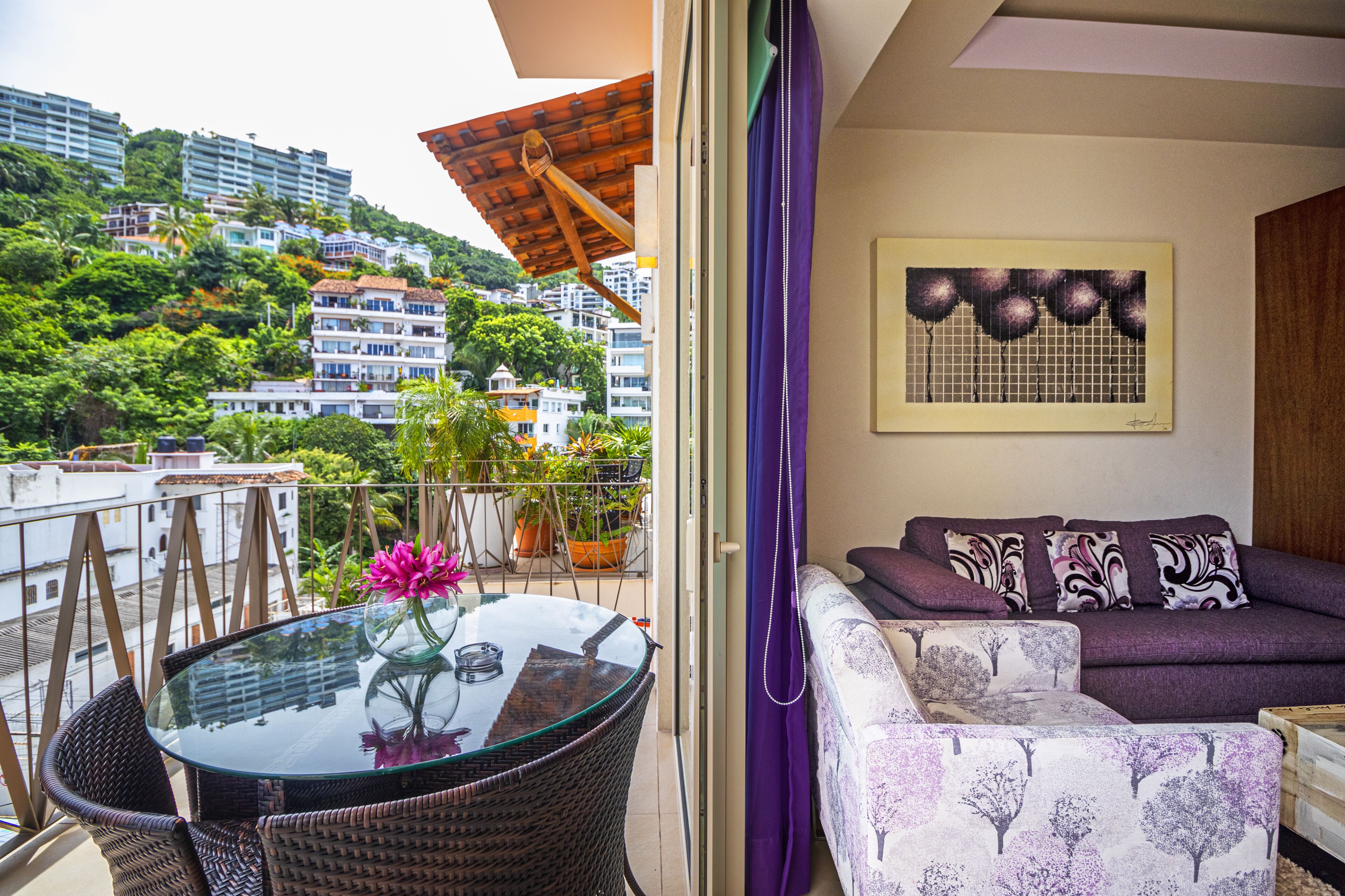 A balcony with a glass table, wicker chairs, a purple sofa, and a view of a hillside with buildings.