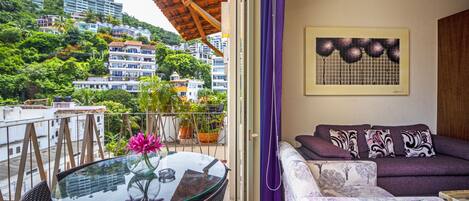 A balcony with a glass table, wicker chairs, a purple sofa, and a view of a hillside with buildings.