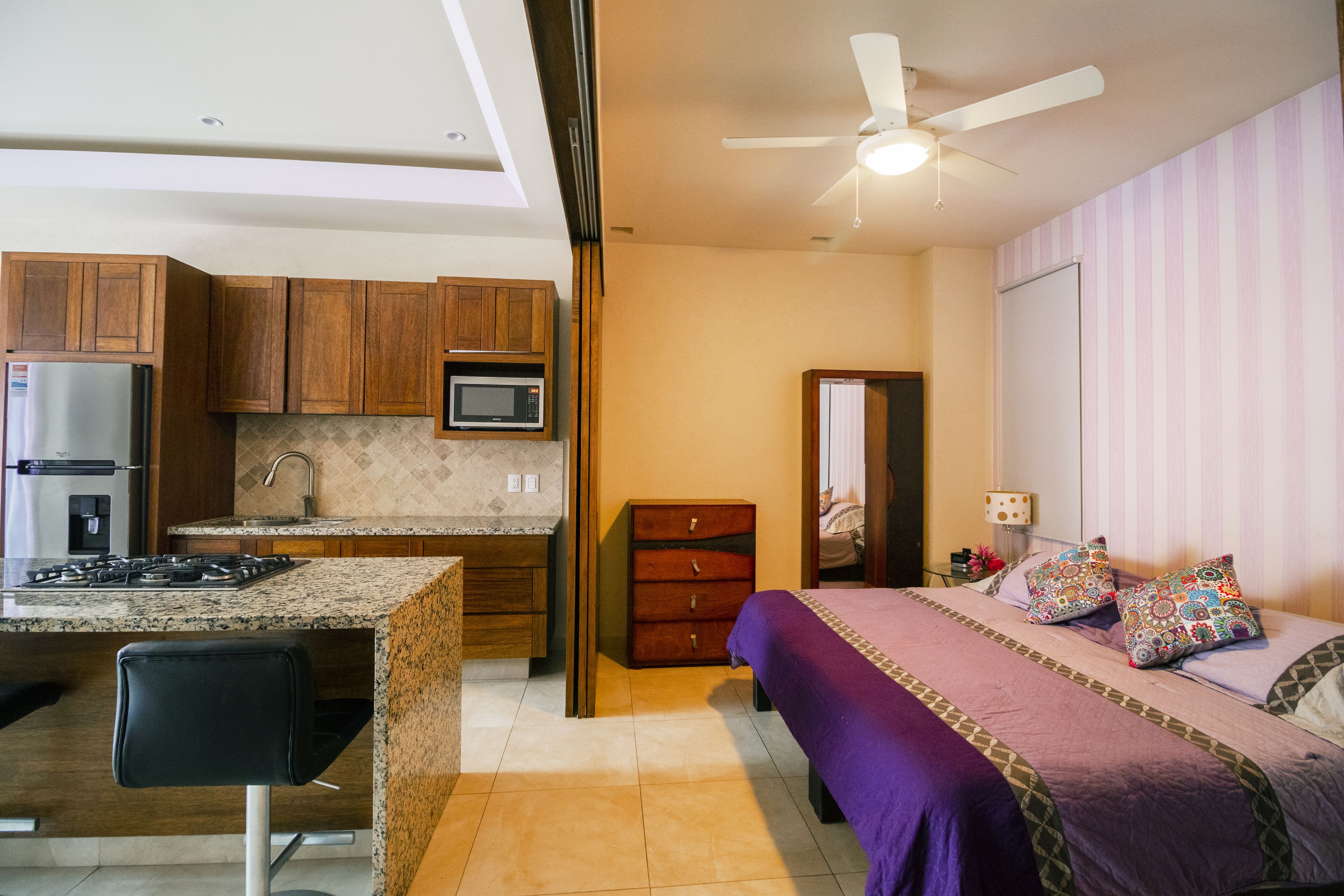 Cocina compacta con refrigerador, microondas y estufa, junto a un dormitorio con cama, almohadas y mesita de noche.