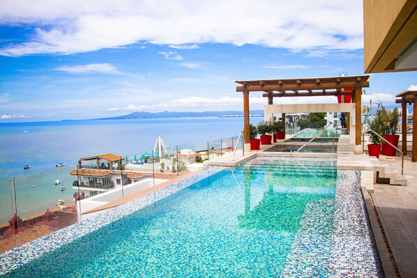 Una piscina con vistas al mar y las montañas, una pérgola y macetas rojas.