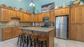 A kitchen with wooden cabinets, a granite countertop, a stainless steel refrigerator, and a central island with stools. - The Tiki (South Padre Island)