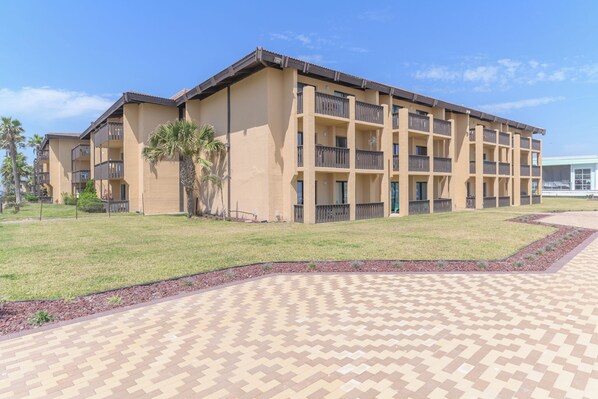 A multi-story hotel with a beige exterior, balconies on each floor, and a paved courtyard with a patterned walkway. - The Tiki (South Padre Island)