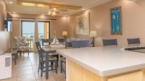 A spacious living area with a large island, a dining table, and a sofa. There is a ceiling fan, a large painting, and a view of the ocean through the windows. - The Tiki (South Padre Island)
