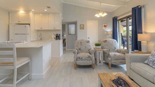 A modern kitchen with white cabinets, a built-in refrigerator, and a central island. There are two armchairs with a patterned upholstery, a wooden coffee table, and a sofa with a cushion. A chandelier hangs from the ceiling, and a window with blue curtains is visible. - The Tiki (South Padre Island)