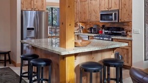 Uma cozinha com armários de madeira, eletrodomésticos de aço inoxidável, bancada de granito e quatro banquetas.