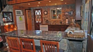 Una cocina con encimera de granito, sillas de madera, pared de piedra y un refrigerador.