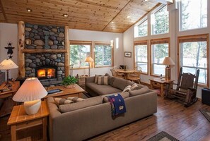 A cozy living room with a stone fireplace, wooden ceiling, and large windows offering a view of the outdoors. - Cristallina Greens (Truckee)