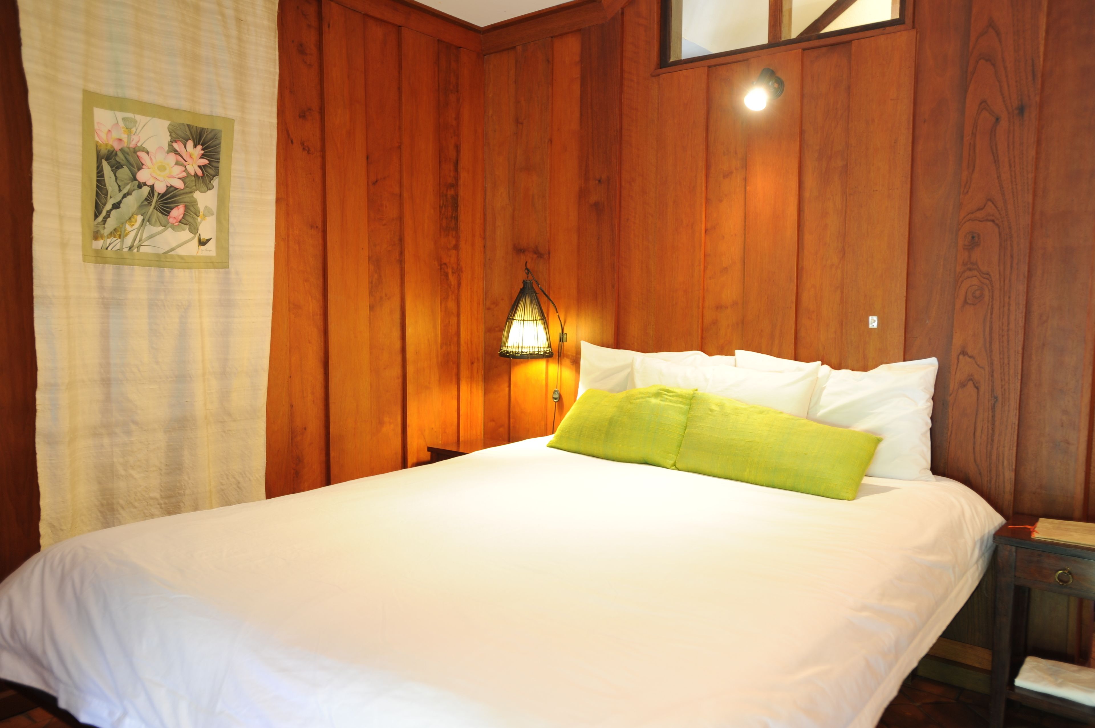 A neatly made bed with white and green pillows in a room with wooden paneling and a nightstand with a lamp.