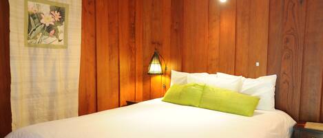 A neatly made bed with white and green pillows in a room with wooden paneling and a nightstand with a lamp.