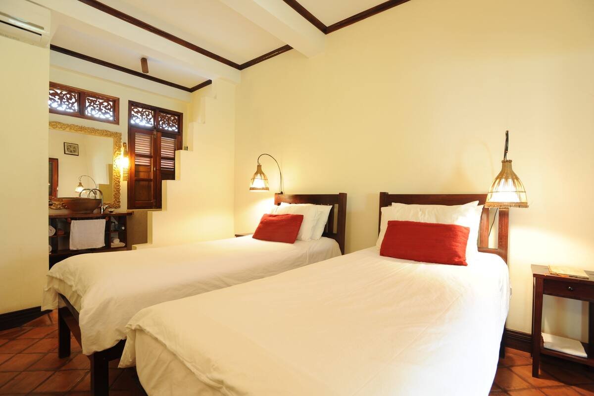 Une chambre d’hôtel avec deux lits, une tête de lit en bois, une table de chevet, une lampe et une fenêtre agrémentée de volets décoratifs.
