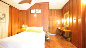 A hotel room with wooden paneling, a bed with white linens, a nightstand, a small table, a mirror with lights, and a bathroom with a sink and mirror.