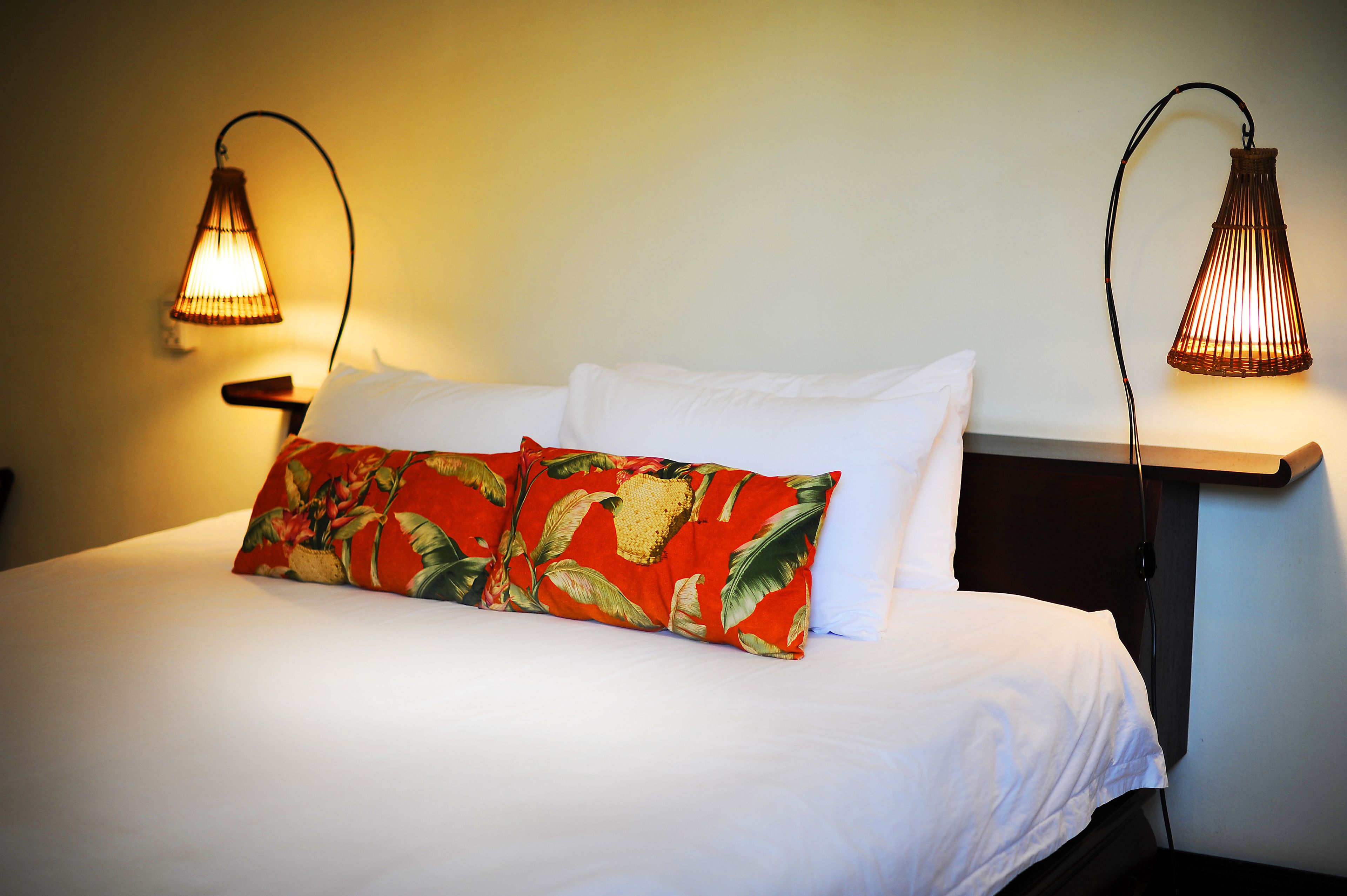 A neatly made bed with white linens and a single red and green floral pillow. Two wall-mounted lamps provide lighting.