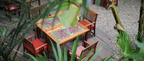 A dining area with wooden tables and chairs, surrounded by greenery.