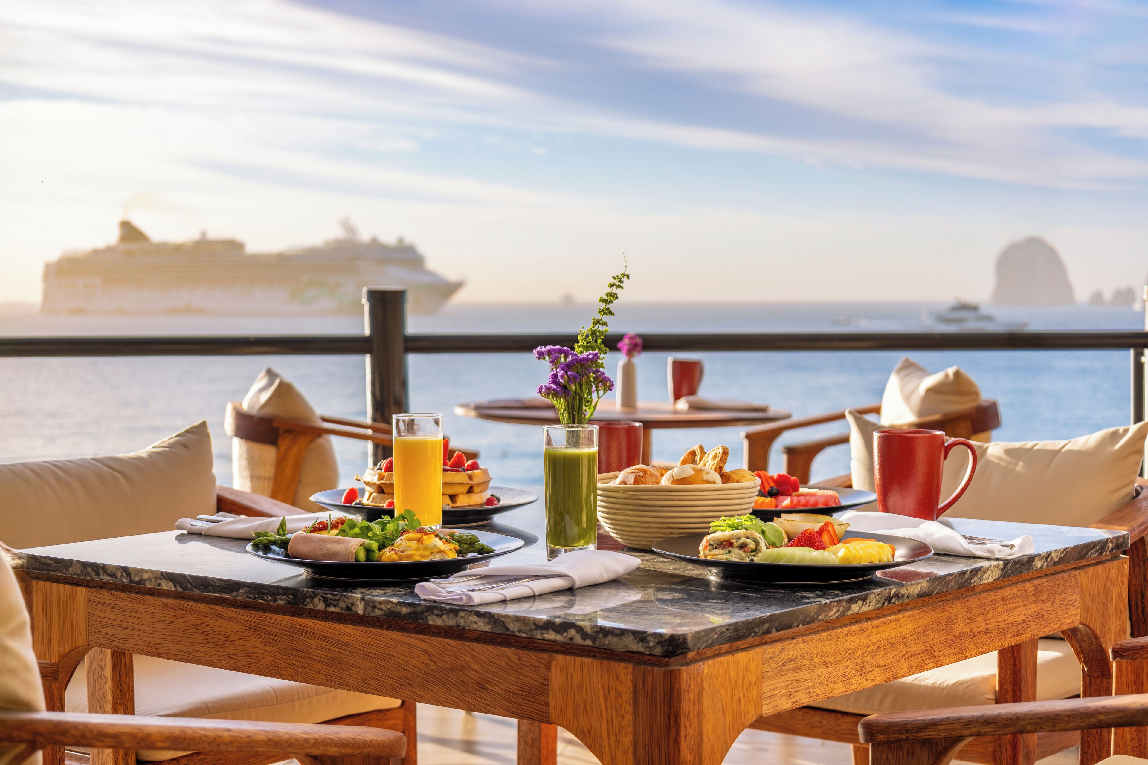 Ein Esstisch mit Frühstücksangeboten, einem Getränk und einer Blumenvase, mit Blick auf ein Gewässer, in der Ferne ein Kreuzfahrtschiff.