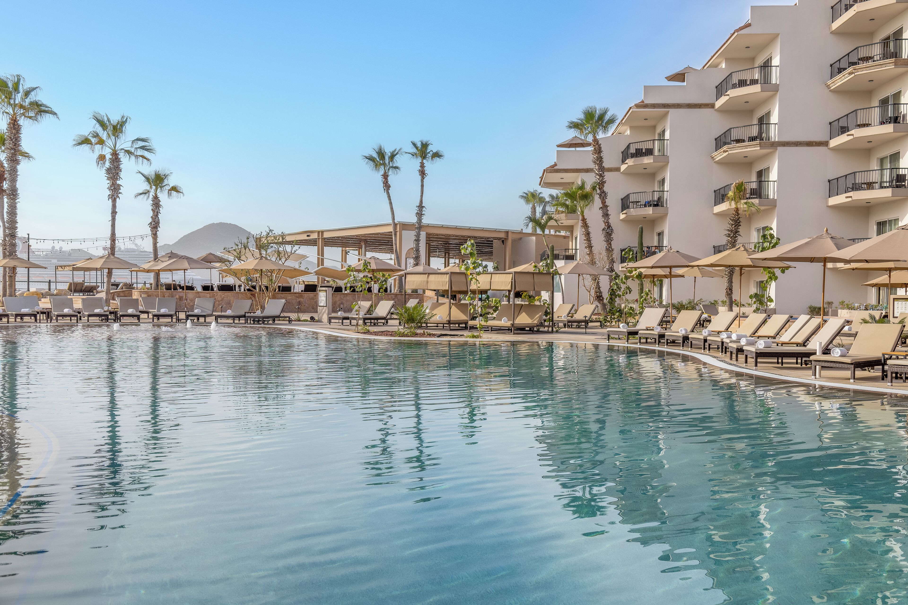 A poolside area with lounge chairs and umbrellas, a building with balconies, and palm trees.