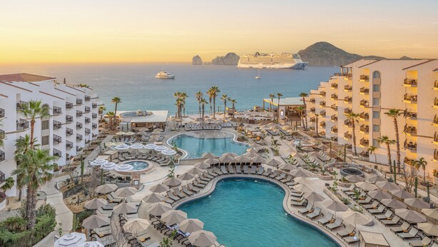 Een resort met meerdere zwembaden, ligbedden en een cruiseschip in de verte.