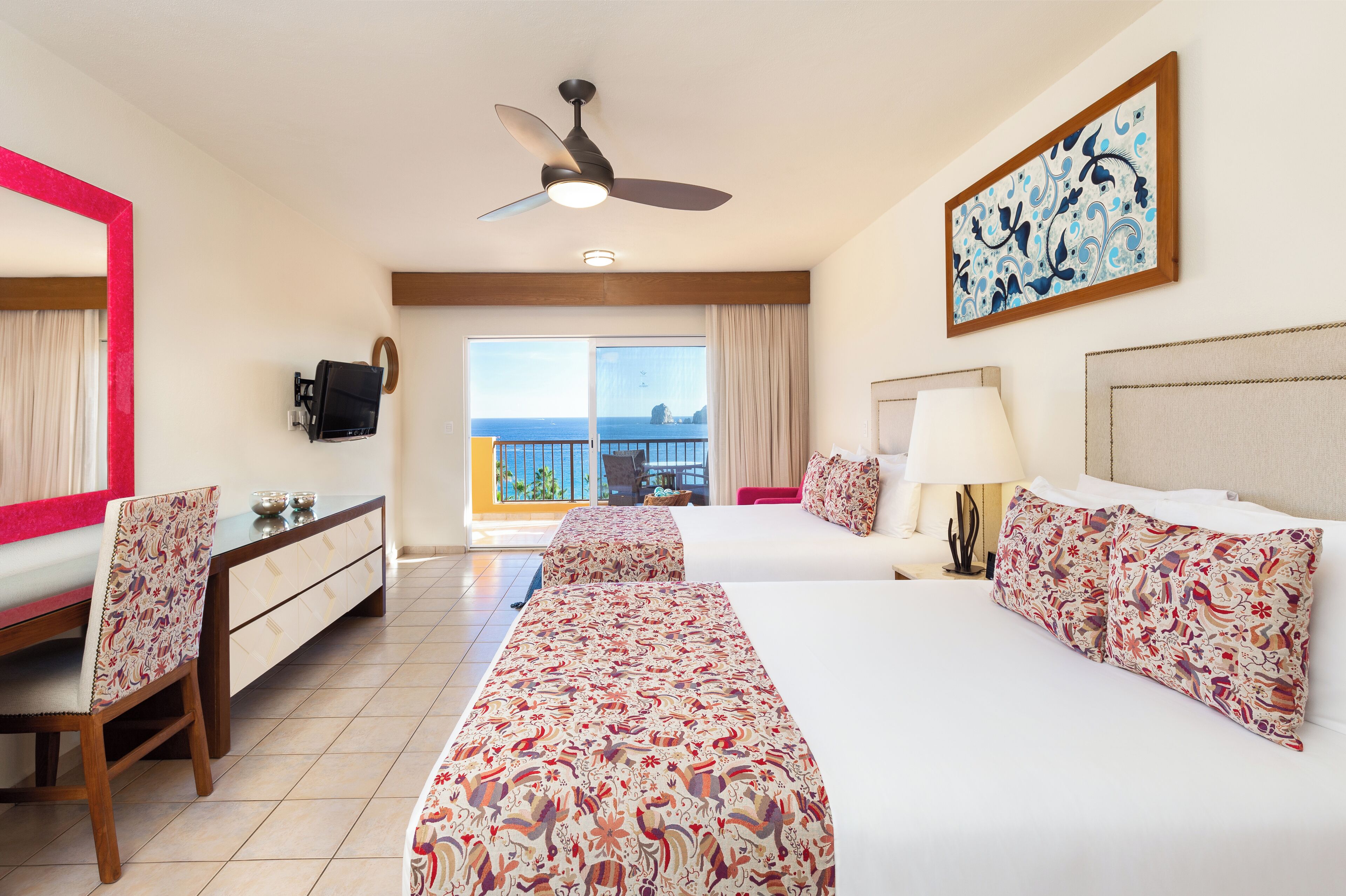 A hotel room with two beds, a desk, a chair, a ceiling fan, a TV, a balcony with a view, and a painting on the wall.
