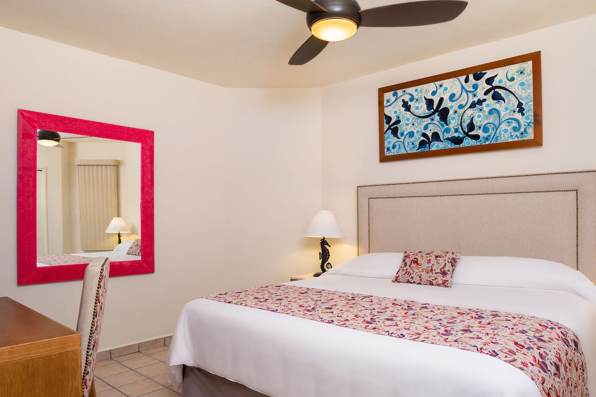 A hotel room with a bed, a desk, a chair, a ceiling fan, a wall-mounted lamp, and a painting.
