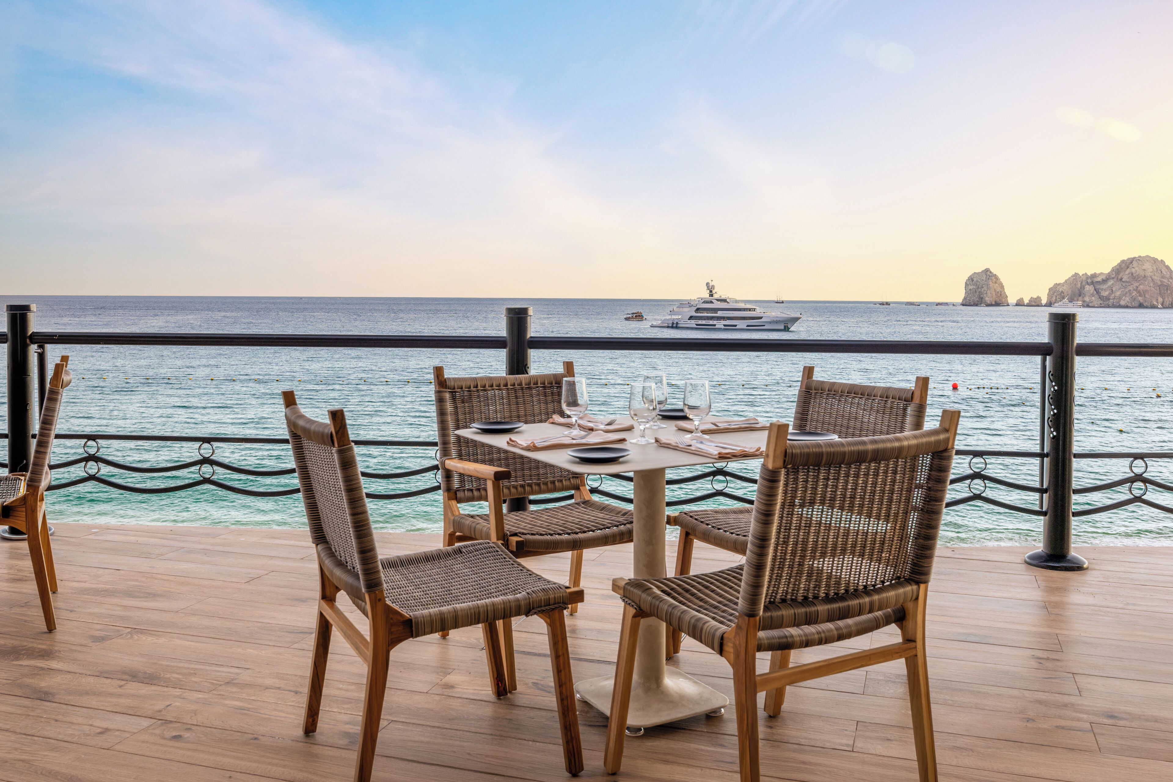 Una terraza con sillas de mimbre y una mesa preparada para comer, con vista a un mar tranquilo y un yate a lo lejos.