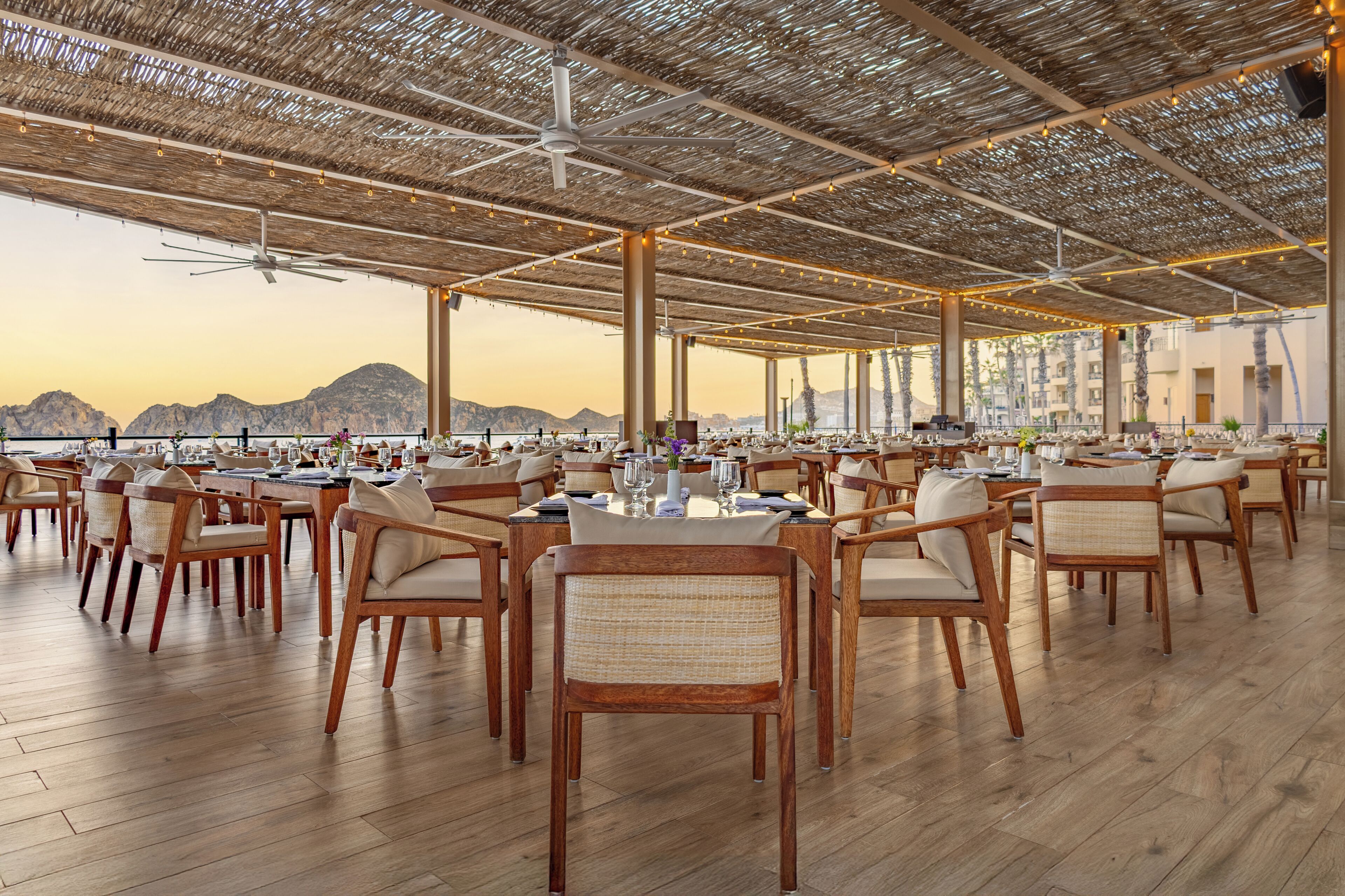 A spacious outdoor dining area with multiple tables and chairs, a thatched roof, and a view of distant mountains.