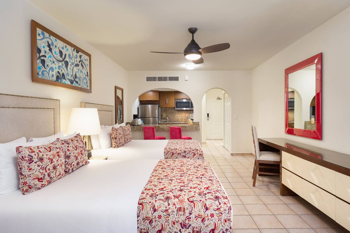 A hotel room with a bed, a desk, a chair, a ceiling fan, a kitchen area, and a large mirror.