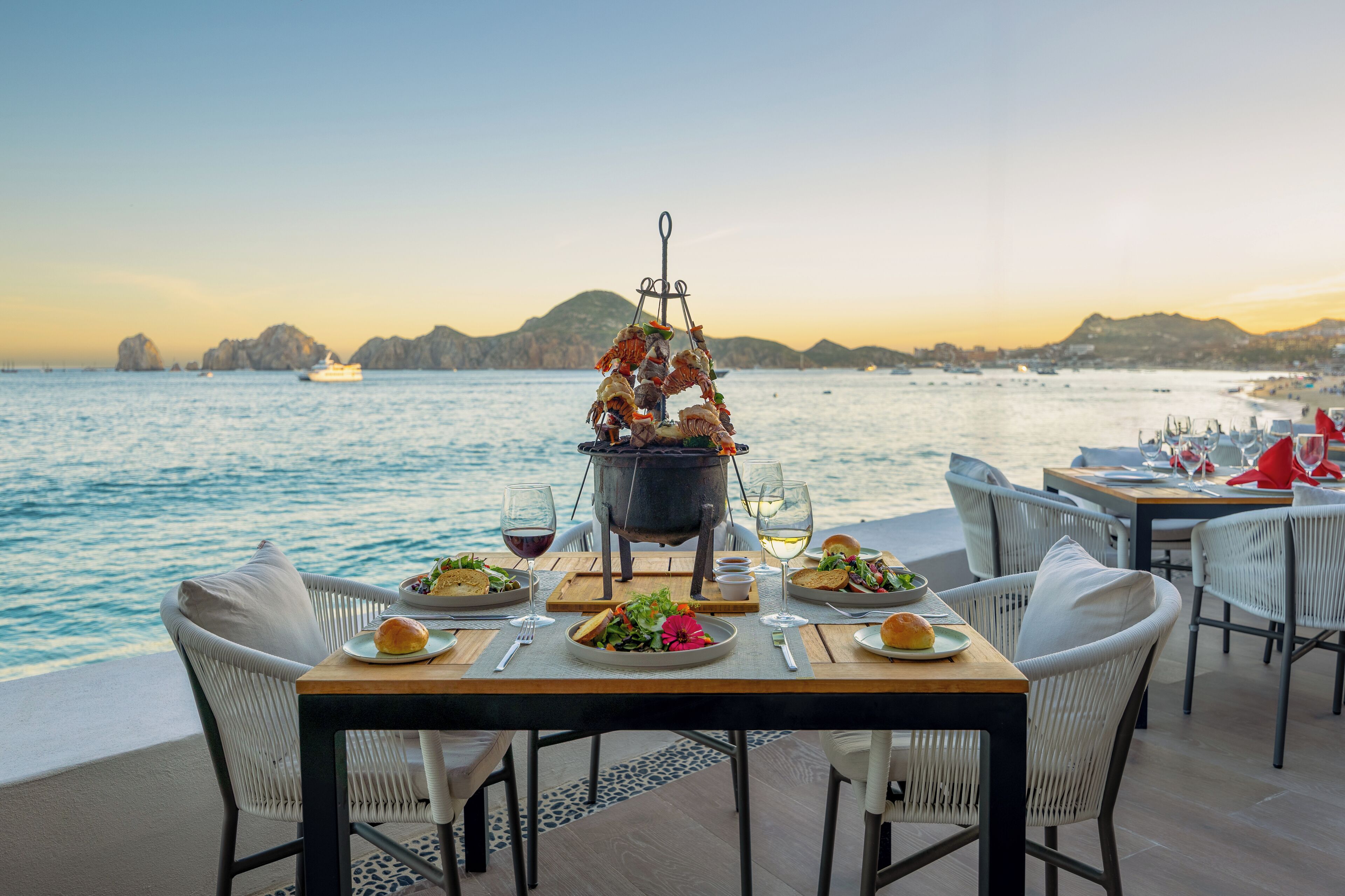 An outdoor dining area with a view of the sea, a table set with plates, glasses, and a central cooking station.