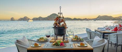 An outdoor dining area with a view of the sea, a table set with plates, glasses, and a central cooking station.