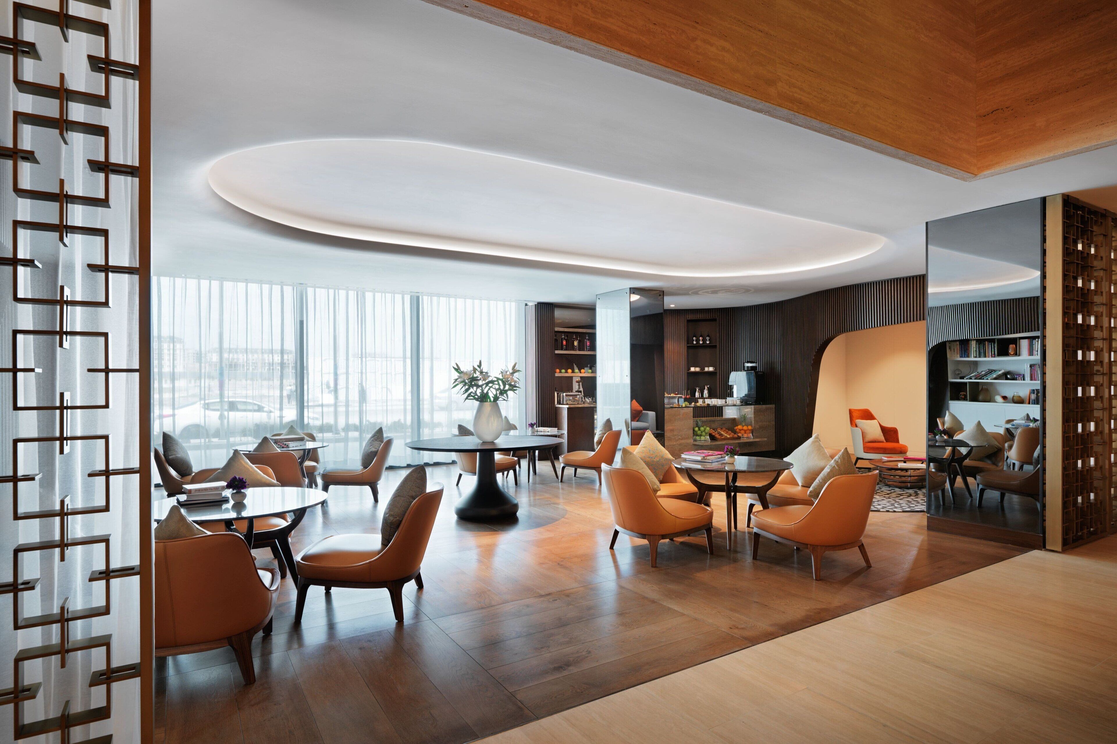 A modern hotel lobby with a wooden floor, orange chairs, a round table, and a large window offering a city view.