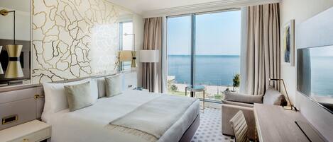 A hotel room with a large bed, a desk, a chair, a TV, and a view of the sea.
