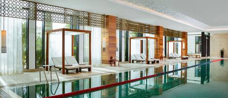 A modern indoor swimming pool with clear water, sun loungers, and a view of greenery outside.