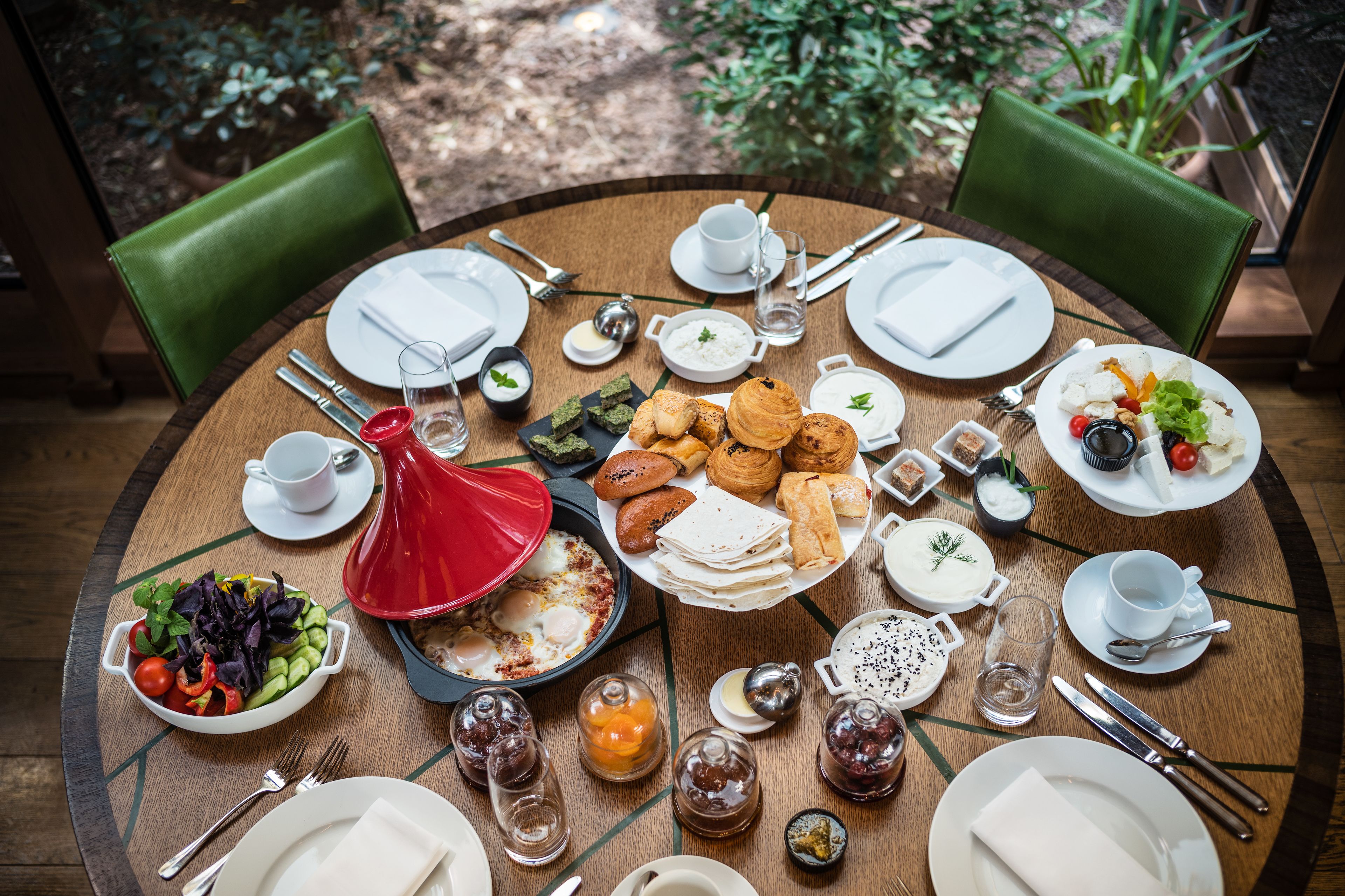 A round dining table with various breakfast items, including eggs, pastries, and salads, surrounded by green chairs.