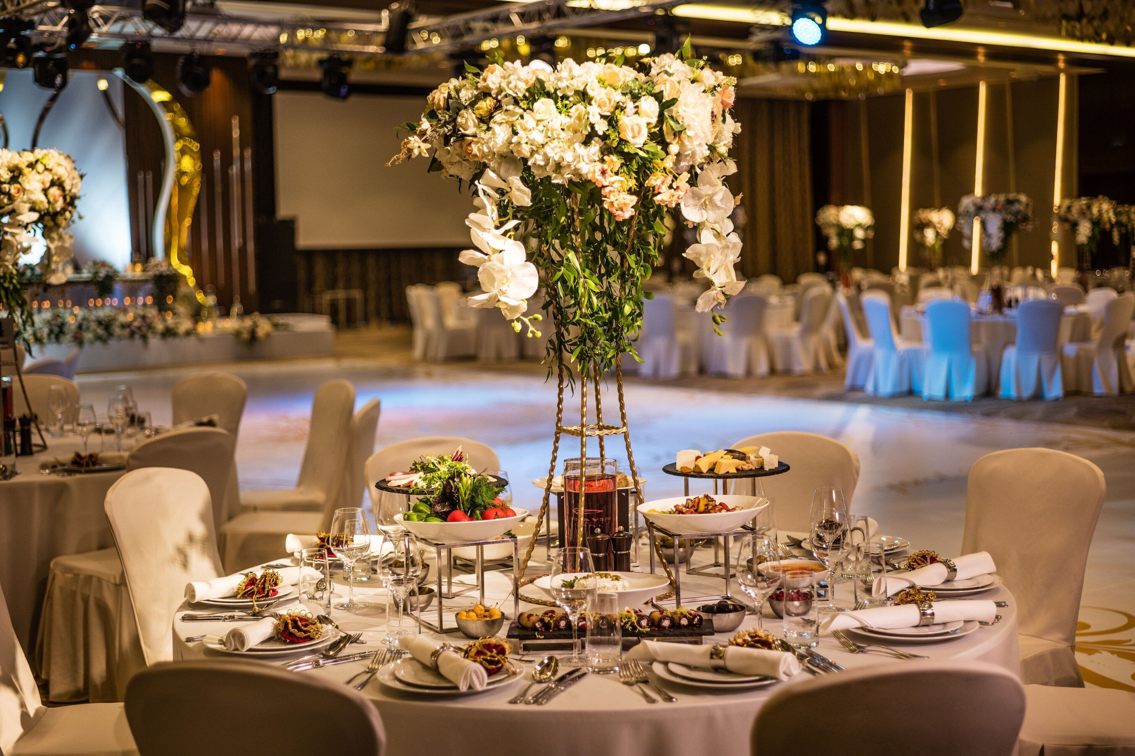 A banquet hall with round tables set for a formal event, featuring floral centerpieces and a tiered stand.