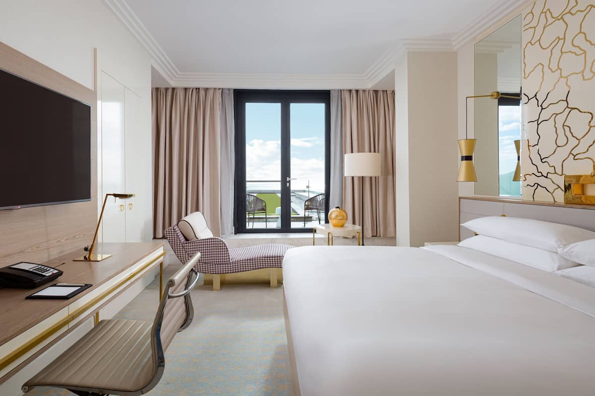 Habitación de hotel con cama, escritorio, silla y un ventanal con cortinas.