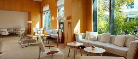 A modern hotel lobby with a large sofa, armchairs, and tables. Large windows offer a view of greenery outside.