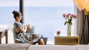 Quarto com uma grande janela com vista para o oceano, um sofá, uma mesinha com um vaso de flores e um abajur.