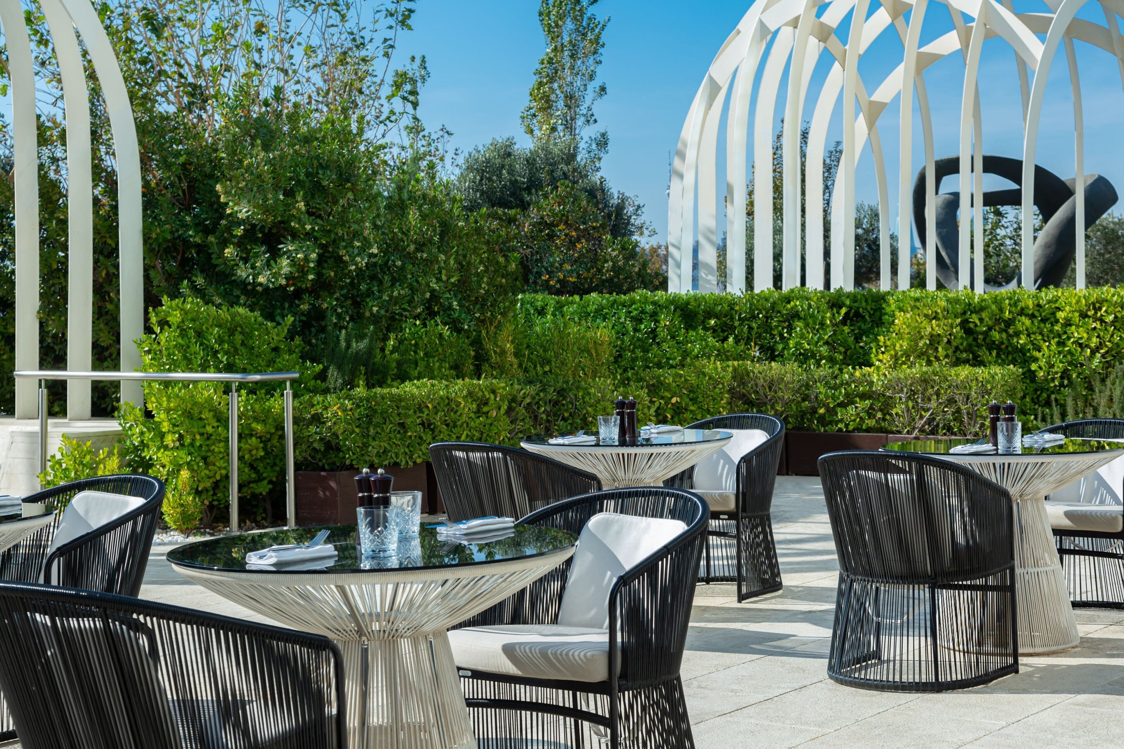 Outdoor seating with round tables and black chairs, surrounded by greenery and a white structure in the background.