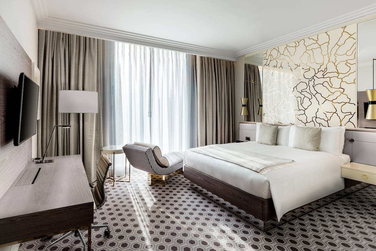 Habitación de hotel con una cama grande, un escritorio con televisor, una silla y una alfombra estampada.